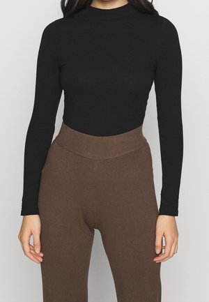 Vrouw die een nauwsluitend zwart topje met lange mouwen draagt, ingestopt in een hooggesneden, geribbelde bruine broek met een V-vormige tailleband.