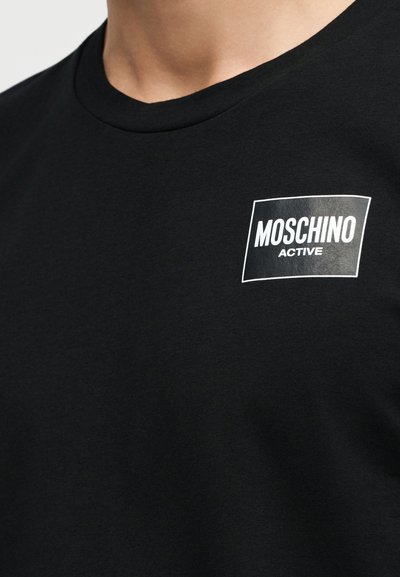 Κοντινό πλάνο ενός ατόμου που φοράει μαύρο μπλουζάκι με μαύρο τετράγωνο λογότυπο με λευκή περίγραμμα και την επιγραφή «MOSCHINO ACTIVE» στο στήθος.
