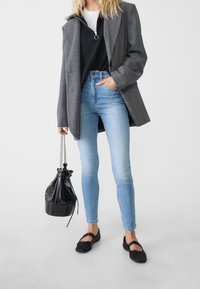 Blazer de cuadros grises sobre un sudadera con cremallera negra, jeans ajustados azul claro y bailarinas negras. Se sostiene una mochila negra con cordones en una mano.