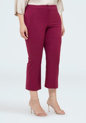 Kvinde iført korte magenta bukser og beige højhælede sandaler, stående mod en ensfarvet baggrund.