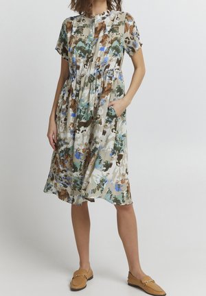 Woman wearing short-sleeve, knee-length floral dress with brown, green, and blue patterns, paired with tan loafers, hand in pocket.