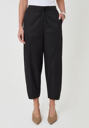 Mujer que lleva pantalones negros anchos recortados con cintura ajustable, tacones marrones de punta afilada y una camiseta blanca.