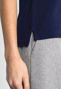 Primo piano della mano di una persona accanto all'orlo di una camicia blu navy con spacco laterale su pantaloni di lana grigia chiara.