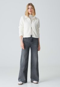 Weiße, flauschige Zip-Jacke mit hohem Kragen, getragen über einem hellen Rollkragenpulli, kombiniert mit hochtaillierten, weiten grauen Jeans und weißen Turnschuhen.