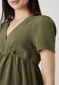 Oliwkowa sukienka z dekoltem w serek, krótkimi rękawami, marszczoną talią i teksturowanym materiałem, charakteryzująca się luźnym krojem i miękkim układem.
