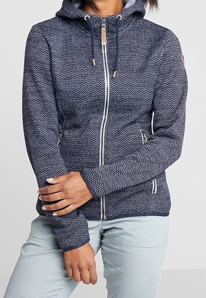 Vrouw draagt een marineblauwe gemêleerde zip-up hoodie met bindkoordjes en lichtgrijze broek tegen een grijze achtergrond.