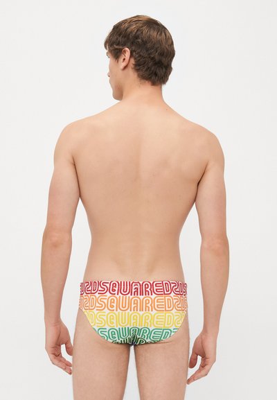 Mann steht mit dem Rücken zur Kamera und trägt mehrfarbige Dsquared2 Badehose mit wiederholtem Logo-Muster auf weißem Hintergrund.