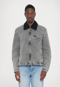Veste en denim grise avec col en velours côtelé noir, fermeture éclair à l'avant et poche poitrine. Texture légèrement usée, assortie à un jean bleu clair.