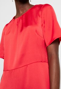 Femme portant un haut en satin rouge vif à manches courtes avec un col rond et de subtils détails de couture à l'avant, sur un fond léger uni.