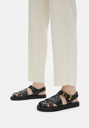 Černé kožené sandály s překříženými pásky a výraznou zlatou sponou. Strukturovaný povrch a plochá gumová podrážka. Nošeny s kalhotami světlé barvy.