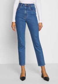 Džíny z modrého denimu s vysokým pasem a rovnými nohavicemi, které mají pět kapes a klasické zapínání na knoflík, doplněné černými lodičkami.