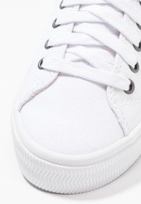 Nahaufnahme der Spitze eines weißen Leinenschuhs mit weißen Schnürsenkeln, die durch Metallösen gezogen sind, und einer strukturierten Gummisohle.