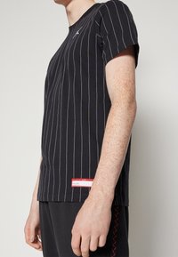 Hombre vestido con una camiseta negra a rayas con un pequeño logo de Jumpman en blanco y una etiqueta rectangular roja cerca del dobladillo, de pie frente a un fondo liso.
