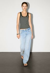Een jonge vrouw loopt zelfbewust, gekleed in een zwart-wit gestreept tanktop en losse blauwe spijkerbroek, met minimale accessoires en blote voeten.