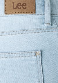 Tejido de mezclilla azul claro con costuras visibles. Presenta una etiqueta de cuero marrón con la marca "Lee" en la cintura. Costuras detalladas en los bolsillos.
