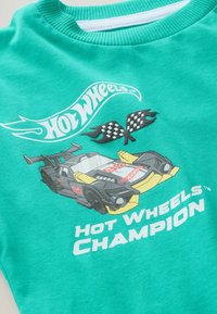 Tirkīza T-krekls ar apaļu izgriezumu, uz kura attēlots sacīkšu automobilis, šaha karogi un balts teksts "Hot Wheels Champion".
