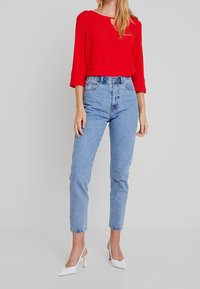 Haut rouge à manches longues avec un col rond, associé à un jean bleu clair taille haute. Le mannequin porte des talons blancs à bout pointu.