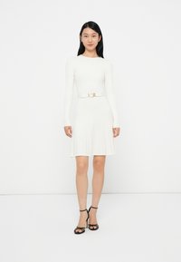 Abito a maglia bianco a coste con maniche lunghe, vita aderente e gonna a trapezio; presenta una cintura con fibbia dorata lucida e sandali neri con tacco alto.