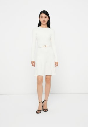 Abito a maglia bianco a coste con maniche lunghe, vita aderente e gonna a trapezio; presenta una cintura con fibbia dorata lucida e sandali neri con tacco alto.