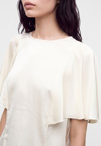 Femme aux cheveux foncés portant un chemisier ample en satin couleur crème avec de larges manches courtes fluides et un col rond.