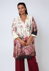 Woman wearing a long cream and pink floral blouse with 3/4 sleeves and burgundy wide-leg trousers, standing against a plain background.