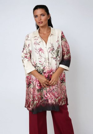 Vrouw in een lange crème en roze bloemen blouse met 3/4 mouwen en bordeauxrode wijde broek, staand tegen een effen achtergrond.