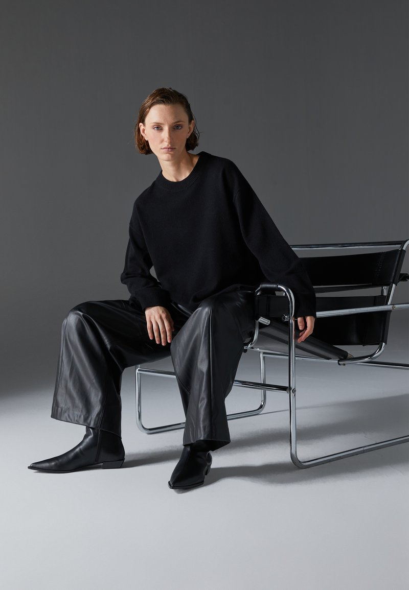 Pull en tricot noir, pantalon large en cuir noir et bottes pointues noires. Assis dans une chaise chromée avec un siège en tissu noir. Fond gris.