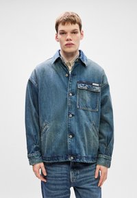 Τζάκετ από τζιν σε ανοιχτό μπλε, διαθέτει μεγάλο γιακά, κλείσιμο με κουμπιά και τσέπη στο στήθος. Η υφή φαίνεται φθαρμένη. Συνδυασμένο με ανοιχτό μπλε τζιν.