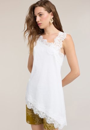 Mujer con cabello largo y ondulado, que lleva un top blanco sin mangas con ribete de encaje y pantalones cortos verde oliva estampados, de pie con las manos detrás de la espalda.