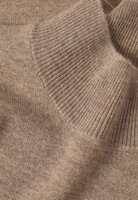 Gros plan sur le tissu beige en maille côtelée d'un pull à col roulé, montrant la texture tricotée et le détail du col replié.