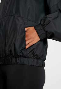 Giacca nera in nylon con polsini e cintura elasticizzati. Presenta tasche laterali e una texture liscia e lucida. Design semplice e sobrio.
