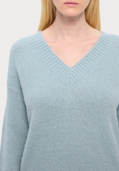 Pulover vâsc de culoare albastru deschis, realizat dintr-un material tricotat moale, cu o textură plăcută, având detalii cu elastic la guler și la manșete.