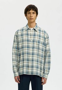 Jeune homme portant une chemise à carreaux bleue et blanche boutonnée sur un t-shirt blanc, associée à un pantalon foncé, debout devant un fond uni.