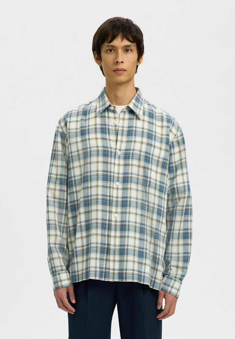 Jeune homme portant une chemise à carreaux bleue et blanche boutonnée sur un t-shirt blanc, associée à un pantalon foncé, debout devant un fond uni.