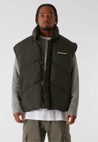 Gepolsterte schwarze Weste mit hohem Kragen und Reißverschluss. Verfügt über diagonale Nähte und ein Markenlabel. Getragen über einem grauen Pullover und einem weißen Hemd.