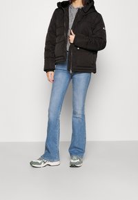 Giacca a vento nera con cappuccio, chiusura con zip e design trapuntato; abbinata a jeans svasati azzurri e scarpe da ginnastica con motivi.