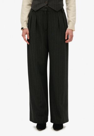 Personne portant un pantalon noir à larges jambes à fines rayures, un gilet noir, un haut crème à manches longues et des chaussures noires, debout sur fond blanc.