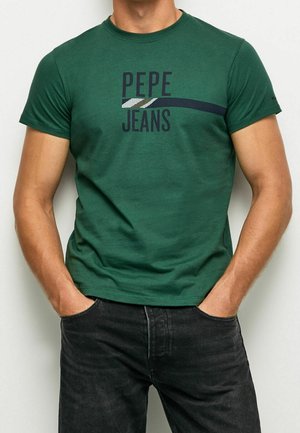 Mand iført en grøn Pepe Jeans T-shirt og sorte jeans med hænderne afslappet i lommerne, overkroppen synlig mod en ensfarvet baggrund.