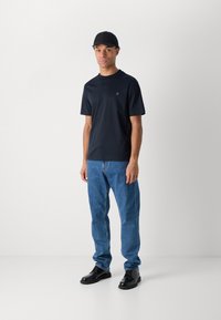 T-shirt à manches courtes bleu marine avec un col rond et un petit logo, associé à un jean bleu clair et des chaussures noires. Design épuré, coupe décontractée.