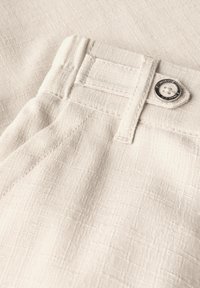 Beige textured fabric with a visible weave pattern; features a waistband with a metal button and stitched detailing.