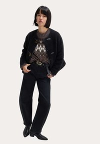 Giacca in pile nera con toggle, maglione a fantasia marrone, pantaloni neri a vita alta e stivaletti neri a punta. Dettaglio della cintura semplice.