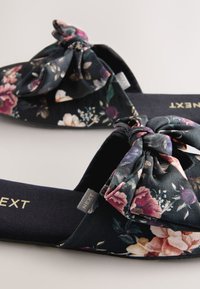 Zapatillas de tela con estampado floral que cuentan con una base negra, un gran lazo decorativo y plantillas acolchadas. La etiqueta dice "NEXT" sobre una superficie.