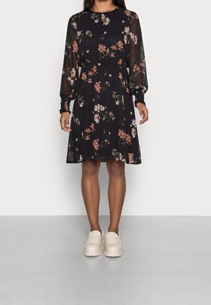 Femme portant une robe noire fleurie longueur genoux avec manches longues transparentes et chaussures blanches à semelle épaisse, debout sur un sol gris.