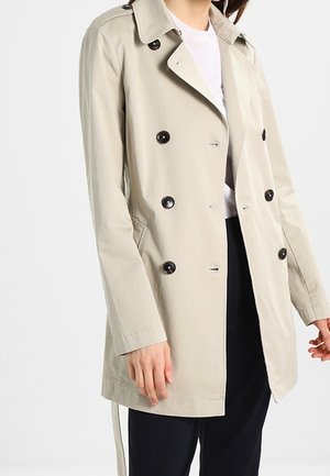 Personne portant un trench-coat beige à double boutonnage avec des boutons noirs sur une chemise blanche et un pantalon noir sur un fond uni.