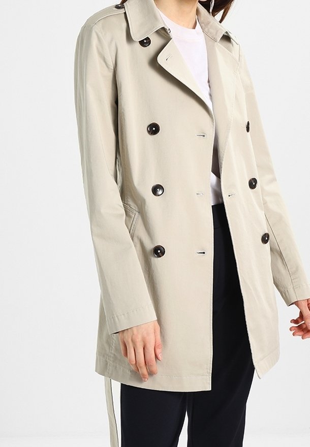Personne portant un trench-coat beige à double boutonnage avec des boutons noirs sur une chemise blanche et un pantalon noir sur un fond uni.