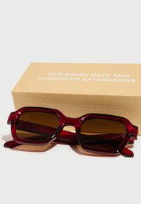 Κόκκινα μεγάλα γυαλιά ηλίου με καφέ φακούς απόχρωσης, τετράγωνο πλαίσιο, γυαλιστερό φινίρισμα, δίπλα σε μια ταμπά χάρτινη συσκευασία με λευκό κείμενο.