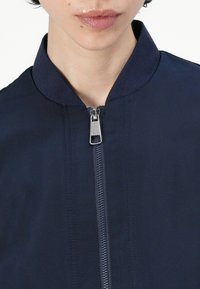 Marineblå zip-jakke med ståkrave, med en glat tekstur og en metallisk lynlås. Synlig syning langs forsiden.