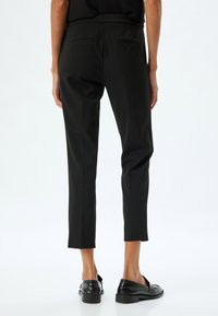 Schwarze, schmal zulaufende Hose aus glattem Material, mit zwei Gesäßtaschen und knöchellanger Passform, kombiniert mit schwarzen, glänzenden Loafers.