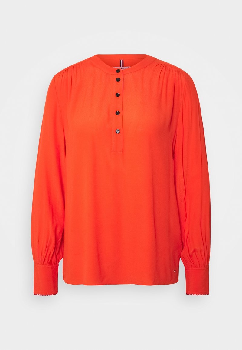 Blouse orange vif à manches longues avec encolure ronde, cinq boutons noirs sur la patte de boutonnage avant, et poignets froncés ornés de dentelle.