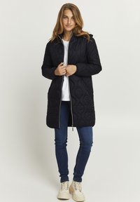 Abrigo negro acolchado con cierre frontal, que cuenta con un cuello alto. Lleva puesto un top blanco y jeans azules, combinado con botas de color crema.